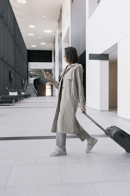 Woman at the airport or congress center with a suitcase is sitting in the waiting area and chatting on her smartphone