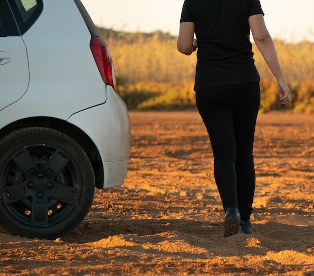 Woman alone dressed in dark clothes walking behind the trunk of a white car at the sunrise