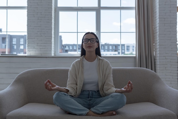 Woman alone at home meditating in front of window sitting on sofa beautiful girl calm down and