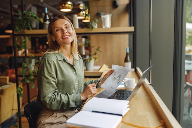 Photo woman analyst working with documents and use laptop while sitting in cozy coworking near window