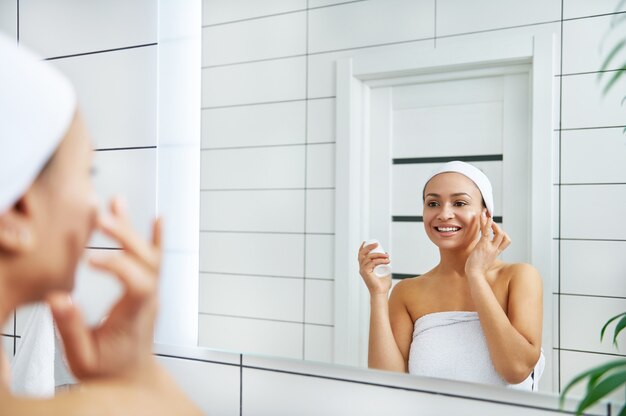 The woman applying cream to face with finger while looking at her reflection in bathroom mirror