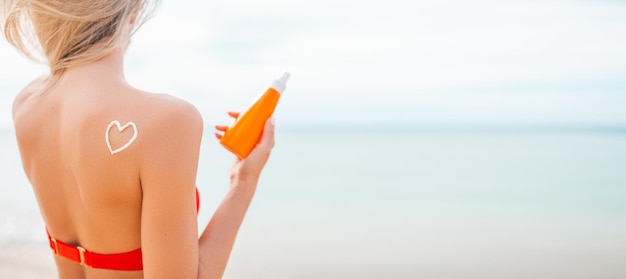 Woman applying sun cream on tanned body In form heart love to protect her skin Girl using sunscreen Female holding suntan lotion moisturizing sunblock on sunny day with blurry sea in the background