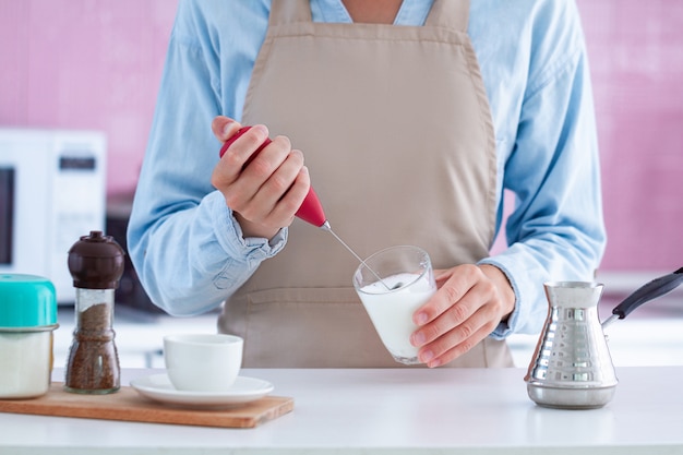 Woman in apron making aromatic coffee with using a milk frother at home