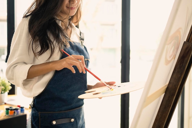 Woman artist holding painting brush and palette painting on canvas at creative workplace