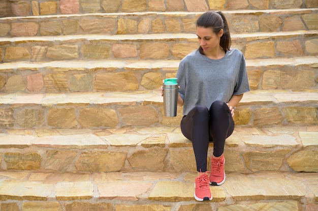 Woman athlete sitting holding aluminum bottle on stairs