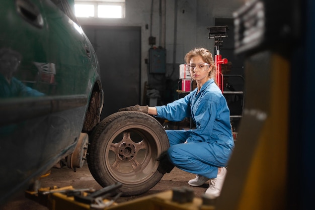 A woman auto mechanic sits near a disassembled car dressed in overalls and safety glasses holds a wheel and looks into the camera