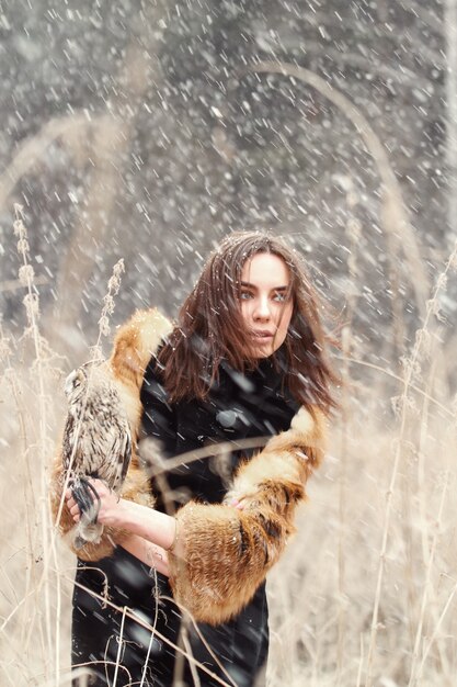 Woman in autumn in fur coat with owl on hand first snow. Beautiful brunette woman with long hair in nature, holding an owl. Romantic, delicate look womans