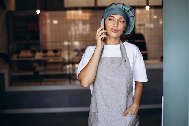 Woman baker in apron standing with phone by the doors at the coffee shop