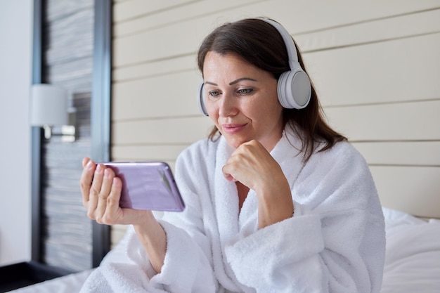 Woman in bathrobe and headphones looking at smartphone screen sitting in bed