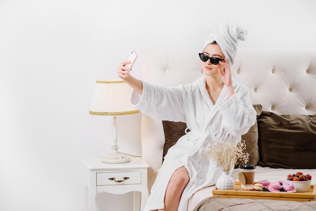 A woman in a bathrobe with towel on her head using smathphone on the bed at home