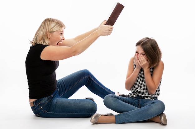 Woman beats teenager daughter with a heavy book. Relationships in the family and the difficulties of home distance learning during the isolation period. White wall.