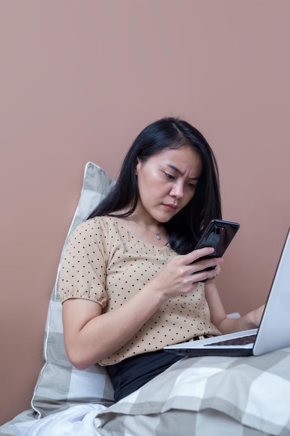 Woman on bed working with laptop while holding cell phone with confused expression