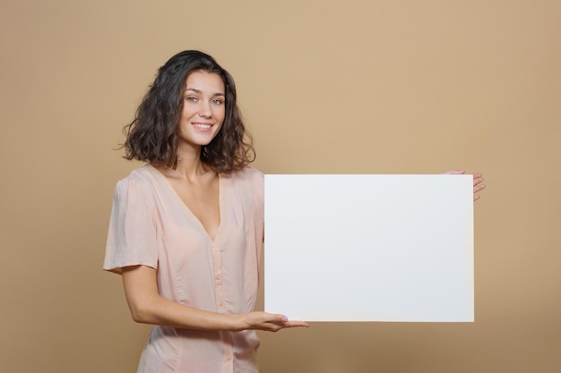 Woman on a beige background smiles and holds a white sign in her hands an advertising poster about