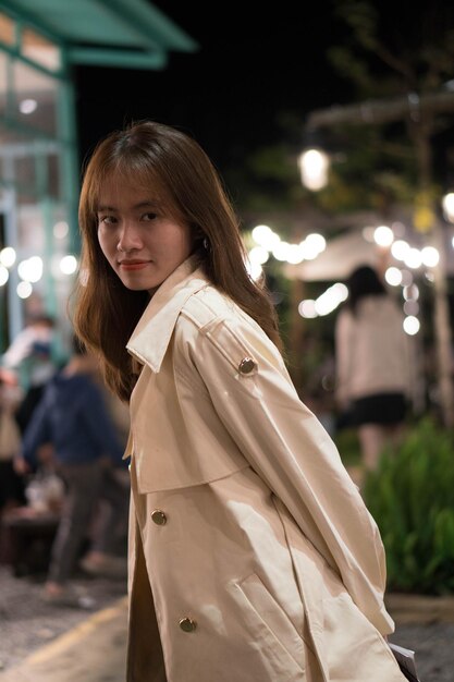 A woman in a beige trench coat stands in front of a street at night.