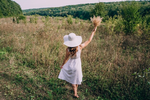 A woman in a big white hat with a bouquet walks among the tall grass. Girl with long hair in a white dress runs in the field. A summer evening.