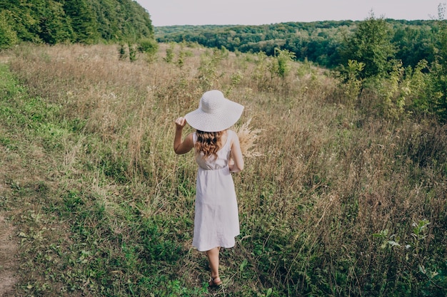 A woman in a big white hat with a bouquet walks among the tall grass