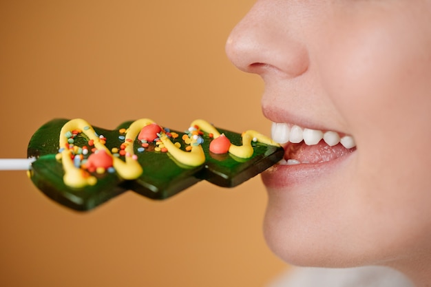 Woman bites with white healthy teeth a sweet lollipop with christmas tree glaze on yellow background