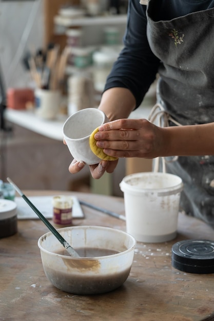 Woman in black apron creates white ceramic mug standing at round wooden table on blurred background