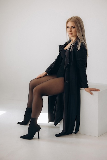 A woman in a black coat sits on a white cube.