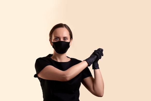 Woman in black dress face mask and gloves joined hands in fists on the side and  over beige wall