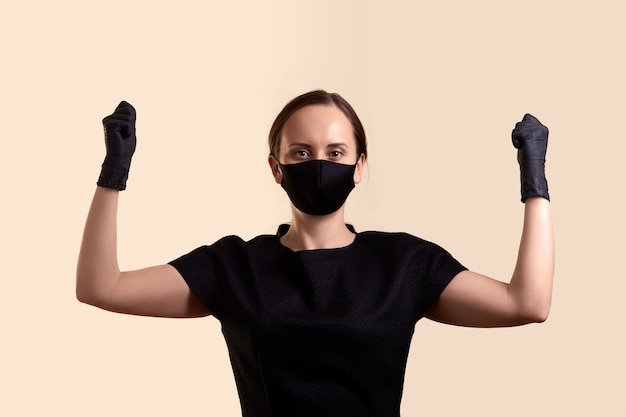 Woman in black dress face mask and gloves raised her hands with fists and  over beige wall