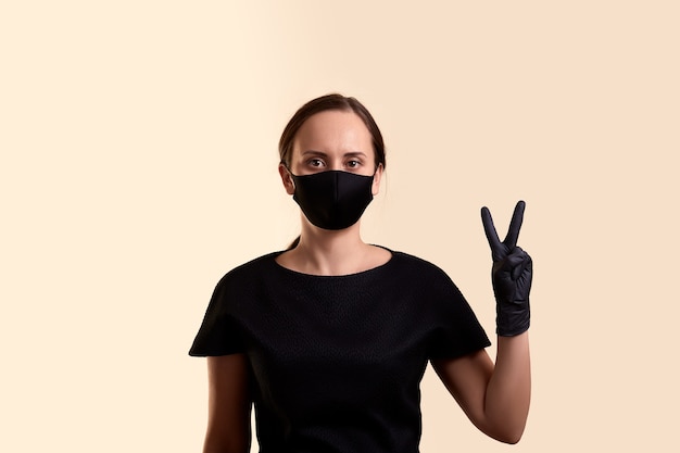 Woman in black dress face mask and gloves showing victory hand and  over beige wall