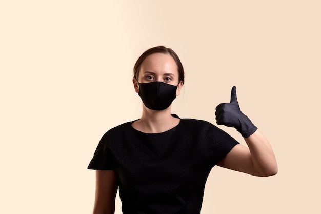 Woman in black dress face mask and gloves showing vthumb up and  over beige wall