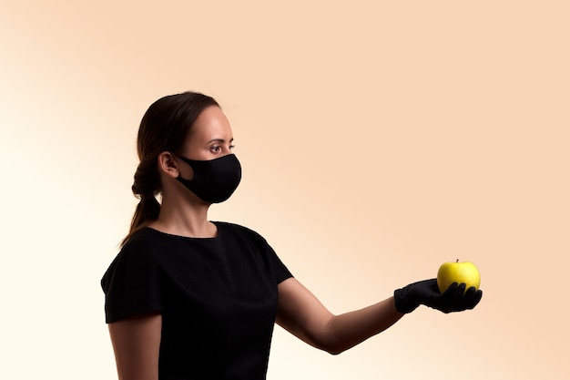 Woman in black dress face mask and latex gloves holding and share red apple over beige wall