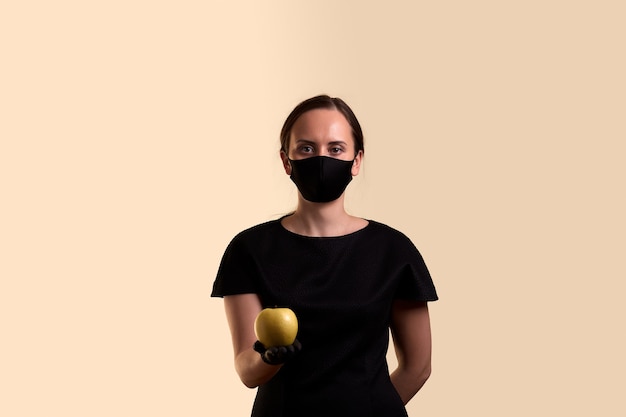 Woman in black dress face mask and latex gloves holding yellow apple and  over beige wall