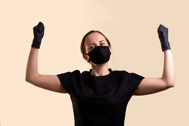 Woman in black dress face mask and latex gloves raised hands with fists bowed her head and  over beige wall