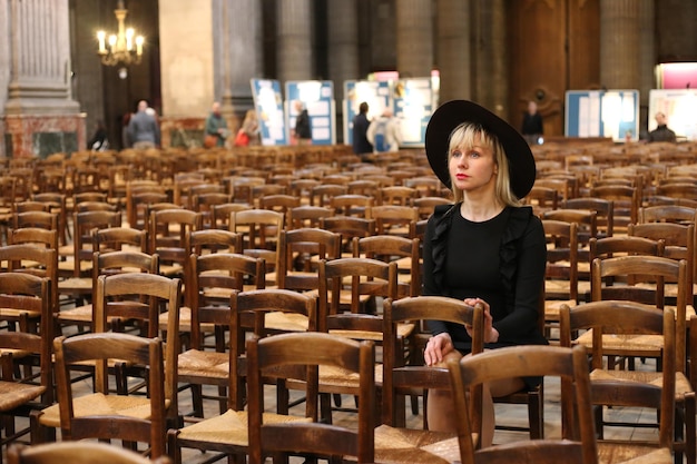 Woman in black dress and hat sits alone in Catholic church. Pray time