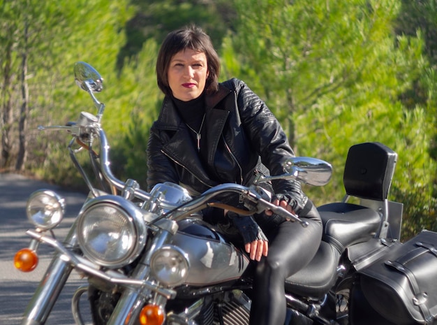 A woman in a black leather biker jacket on a chopper motorcycle in Greece on a road in the forest