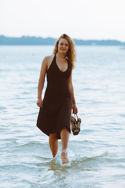 Woman in black light dress walking barefoot by sea beach warm summer evening