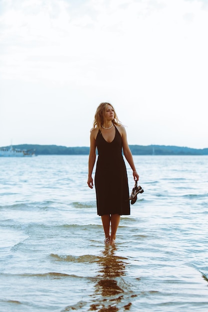 Woman in black light dress walking barefoot by sea beach