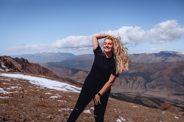 A woman in a black outfit with curly hair smiles sweetly posing against the backdrop of huge mountain hills