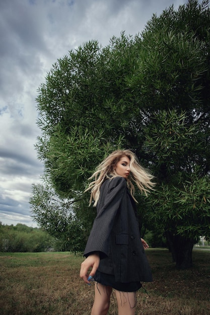 A woman in a black robe and long black shoes walks in front of a tree.