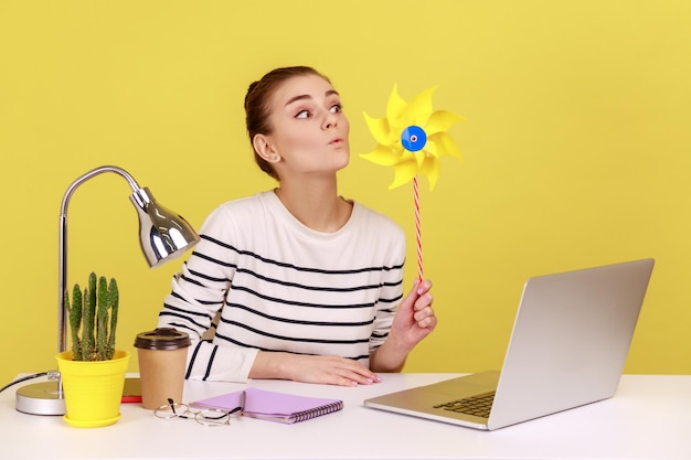 Woman blowing at windmill while sitting on workplace with laptop playing with pinwheel toy on stick