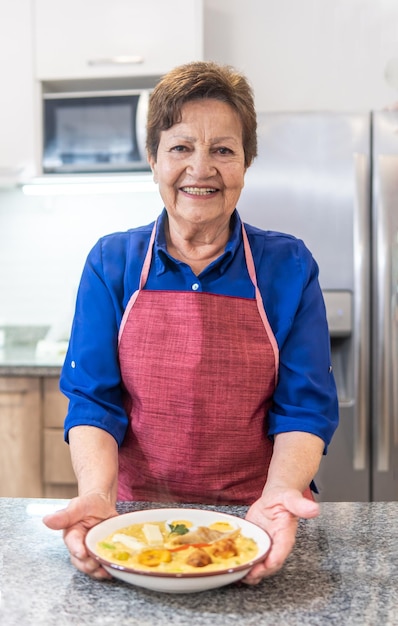 A woman in a blue apron stands in a kitchen, she is holding a piece of pizza.