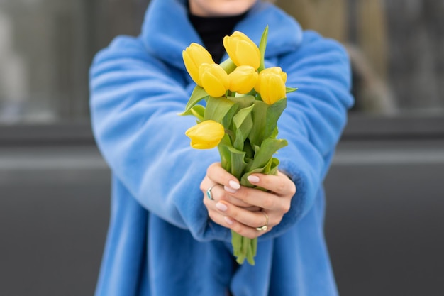 A woman in a blue coat holds a bunch of yellow tulips