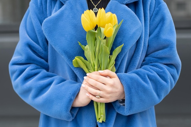 A woman in a blue coat holds a bunch of yellow tulips.