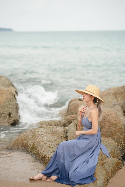 Woman in blue dress and straw hat, sitting on a rock by the ocean.