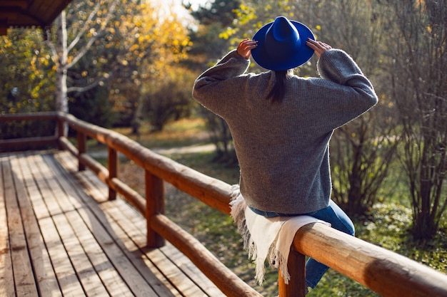 Photo woman in a blue hat and sweater is sitting back on wooden fence near wooden house made of logs in autumn