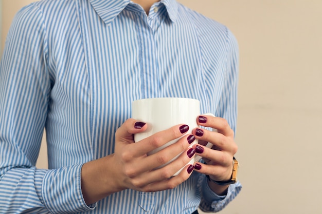 Woman in a blue shirt and red manicure holding a white cup with two hands close up