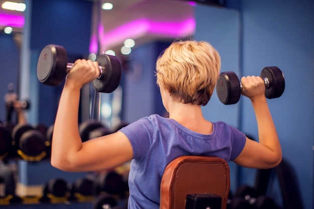 Woman in blue shirt with short hair training with dumbbells in the gym. People, fitness and health concept