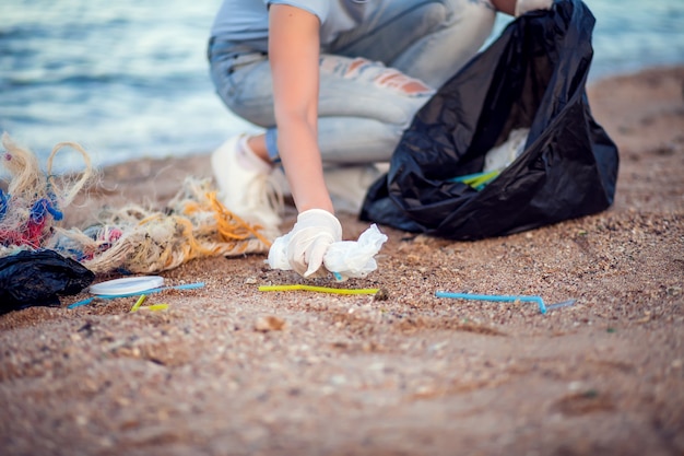 Woman in blue shirt with white gloves and big black package collecting garbage on the beach. Environmental protection and planet pollution concept