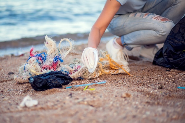 Woman in blue shirt with white gloves and big black package collecting garbage on the beach. Environmental protection and planet pollution concept