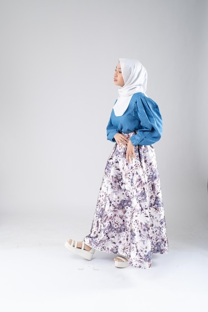 A woman in a blue top and floral skirt stands in front of a white backdrop.