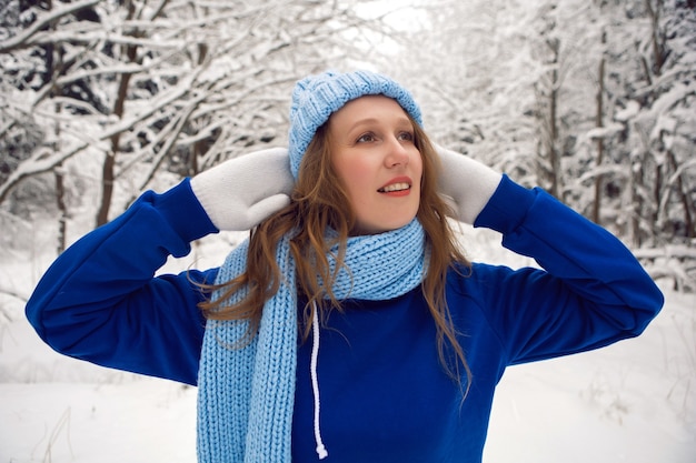 Woman in a blue tracksuit white mittens and scarf stands in winter in a snow covered forest