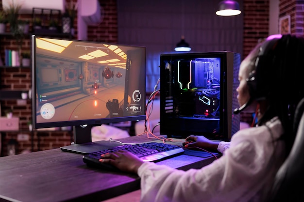 Woman in brick wall living room playing science fiction video game on gaming PC at computer desk, chilling after work. Gamer challenging foes in online multiplayer futuristic shooter