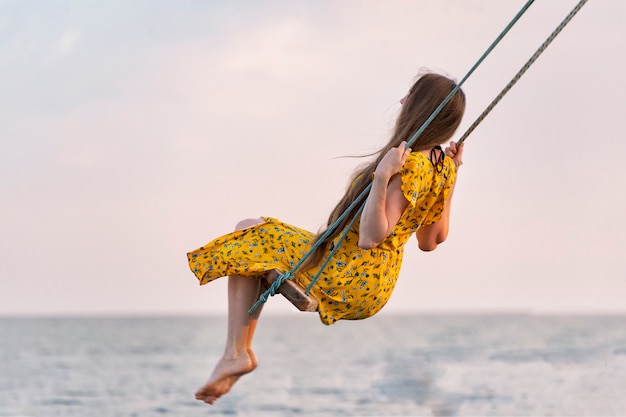 Photo woman in bright yellow dress rides on swing against the sea background. alone with thoughts. loneliness.